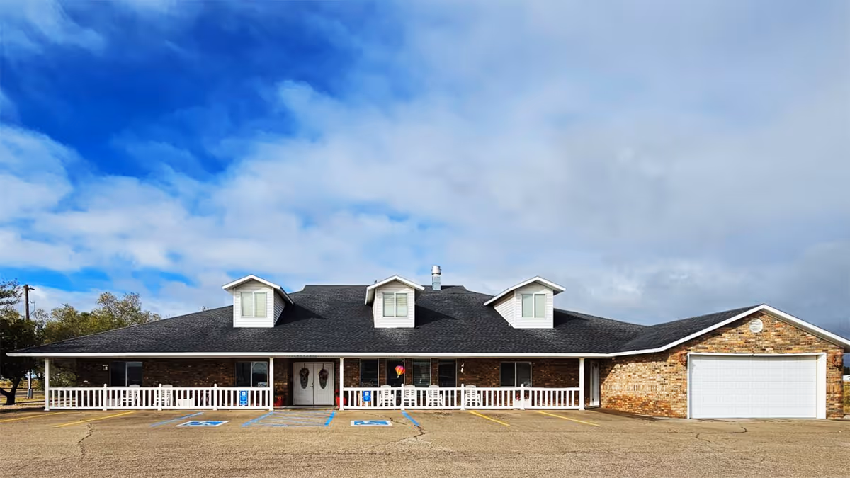 Front exterior view of a single-story brick building with a dark shingled roof featuring three dormer windows. The building has a white railing along the front porch and two double doors at the entrance. There are several parking spaces, including two handicap spots, in front of the building under a partly cloudy blue sky.