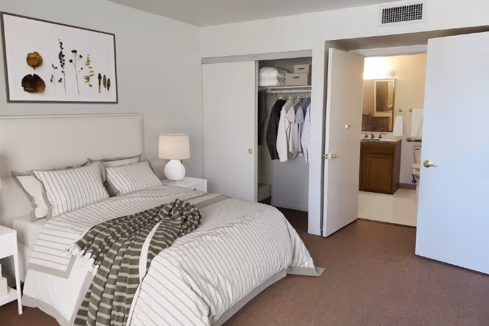A bedroom with a neatly made bed featuring striped bedding and a checkered throw blanket. There is a white nightstand with a lamp on the left side of the bed. Above the bed hangs a framed artwork of dried plants. To the right, there is an open closet with clothes hanging and shelves with storage boxes. Next to the closet is an open door leading to a bathroom with a sink, mirror, and toilet visible.