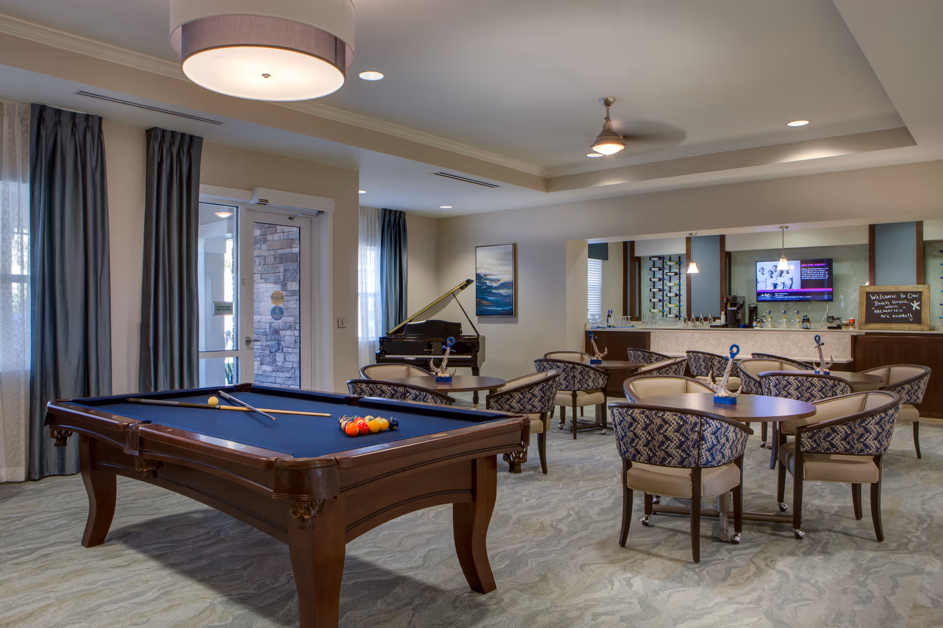 A spacious recreational room in an assisted living facility featuring a pool table with balls and cues, several round tables with patterned chairs, a grand piano in the corner, and a bar area with a television mounted on the wall. The room has large windows with curtains, a ceiling fan, and modern lighting fixtures.