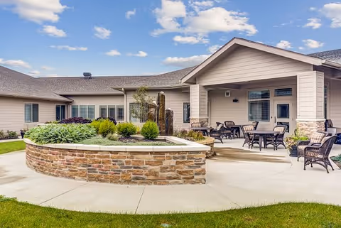Outdoor patio area at Lakewood Memory Care & Assisted Living featuring a circular stone planter with greenery and a water fountain in the center. Surrounding the planter are several wicker chairs and tables under a covered porch attached to a beige building. The sky is partly cloudy with blue patches.