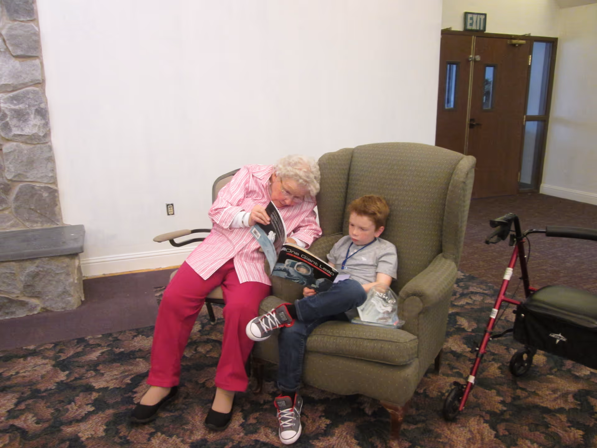 An elderly woman and a young boy sitting together in a green armchair inside a facility. The woman is showing the boy a magazine or book, and they appear to be engaged in reading. There is a red walker nearby and a stone fireplace partially visible on the left side of the image. The setting looks like a common area or lounge with carpeted flooring and an exit door in the background.