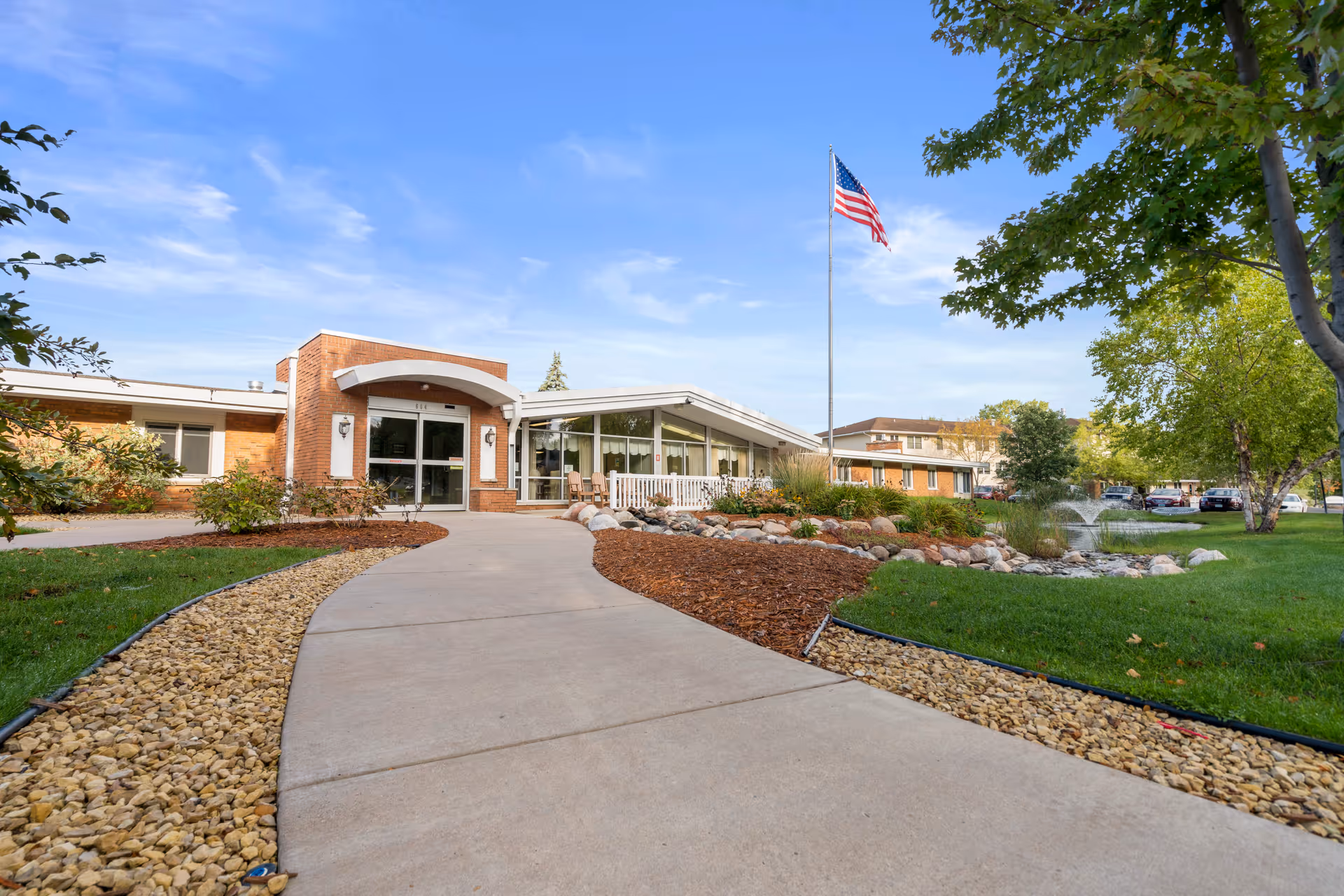 Exterior view of Birchwood Healthcare Center SNF showing a paved walkway leading to the main entrance of a single-story brick building with large windows. There is a well-maintained lawn, landscaping with rocks and mulch, a flagpole with an American flag, and a small water fountain feature. Trees and parked cars are visible in the background under a blue sky with some clouds.
