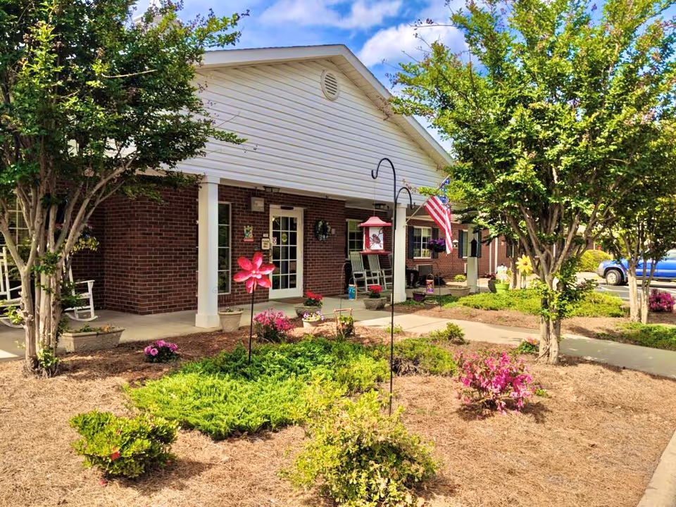 Front exterior view of a brick building with a white porch roof, decorated with potted plants, rocking chairs, an American flag, and garden decorations. There are trees and shrubs in the landscaped area in front of the building, and a blue vehicle is partially visible in the parking area to the right.