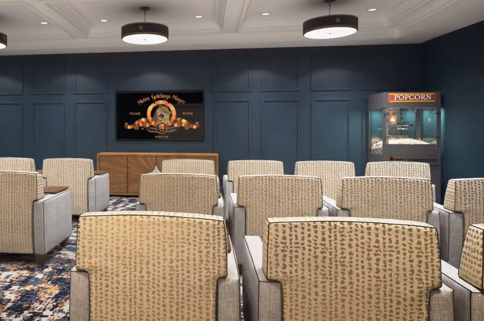 A small theater room with multiple rows of beige patterned armchairs facing a screen mounted on a dark blue paneled wall. To the right of the screen is a popcorn machine. The ceiling has recessed lighting and modern circular light fixtures.
