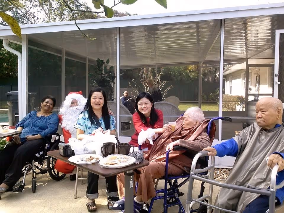 A group of elderly and middle-aged people sitting outdoors near a table with plates and cups. One person is dressed as Santa Claus. Two elderly individuals are in wheelchairs, and one elderly man is using a walker. The setting appears to be a patio area with a screened enclosure and greenery in the background.