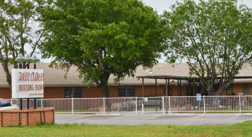 Exterior view of Will-O-Bell Nursing Home building with a brick facade, a white fence in front, several large green trees, and a sign that reads 'Will O'Bell Nursing Home Visitors Welcome'.