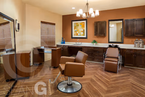 Interior of a salon room with two brown salon chairs, a large mirror on the left, wooden cabinets along the back wall, a countertop with decorative items, and a chandelier hanging from the ceiling. The floor has a herringbone wood pattern.