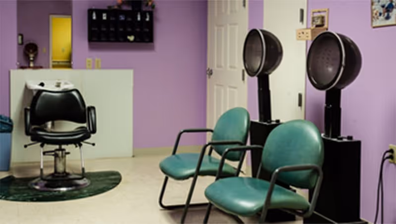 Interior of a salon area with two green chairs in front of two hair dryers and a black salon chair near a white counter against a purple wall.