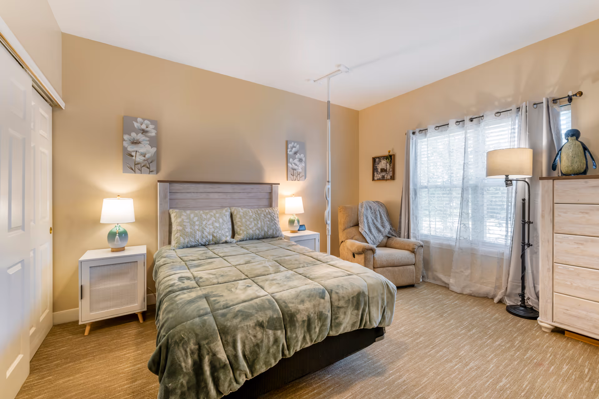A cozy bedroom with a bed covered in a green comforter and matching pillows, two bedside tables each with a lamp, a beige armchair with a blanket draped over it, a tall dresser, a floor lamp, and a window with sheer curtains letting in natural light. The walls are painted a warm beige color and decorated with floral artwork.