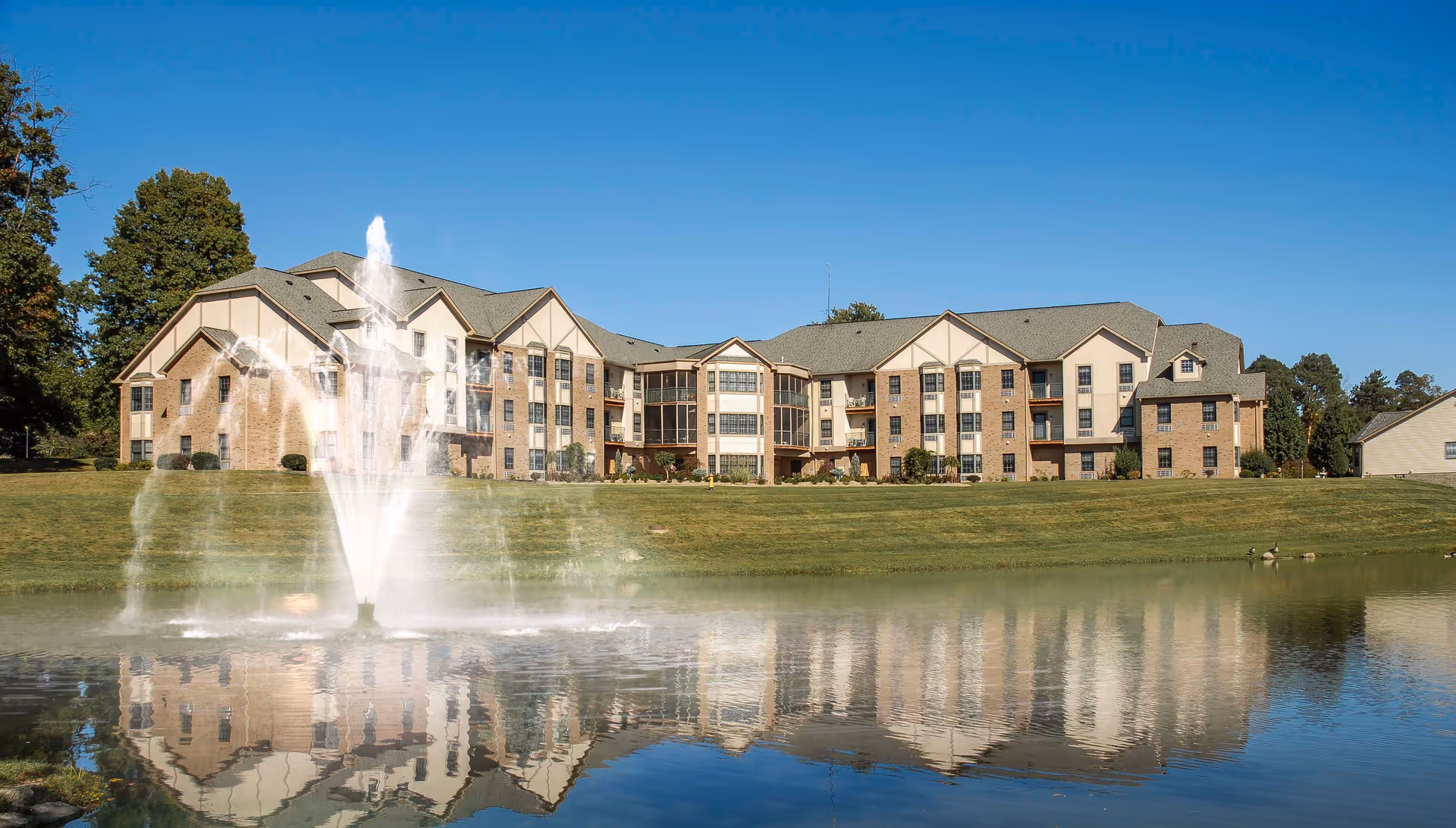 Large multi-story senior living building reflected in a pond with a central fountain and a grassy lawn.