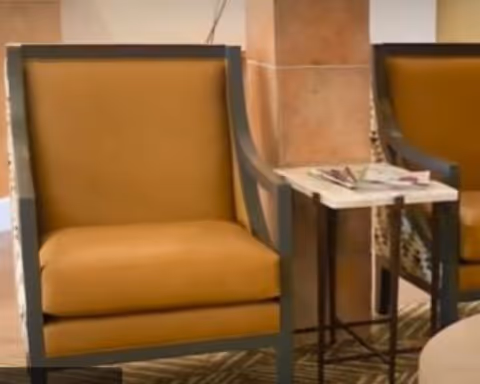 Two mustard yellow cushioned armchairs with dark wooden frames positioned on either side of a small square side table with a white top, placed on a patterned carpeted floor in an indoor setting.