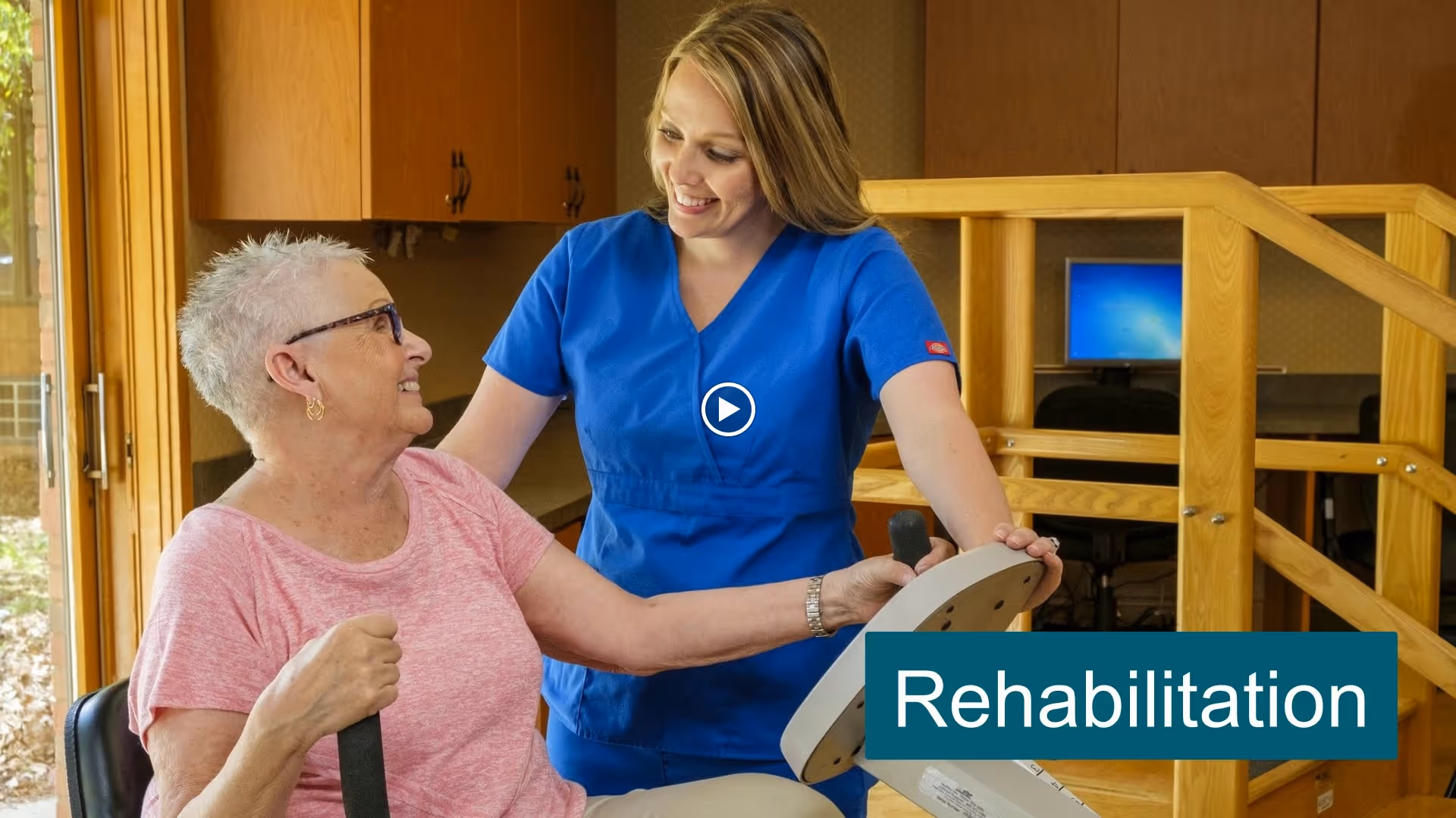 A healthcare worker in blue scrubs assists an elderly woman with short white hair and glasses in using a rehabilitation exercise machine inside a facility room with wooden furniture and a computer in the background.