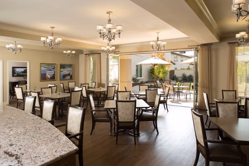 A bright dining room with multiple tables and chairs arranged neatly. The room features chandeliers on the ceiling and large windows with beige curtains allowing natural light to enter. Outside, there is a patio area with tables, chairs, and umbrellas surrounded by greenery.