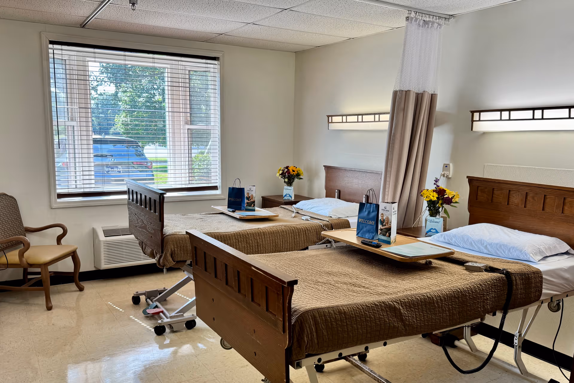 A room with two hospital-style beds with brown bedspreads and white pillows, each equipped with an overbed table holding welcome bags and flowers. There is a wooden chair with armrests near a large window with blinds, and a privacy curtain between the beds. The room has white walls and a tiled floor.