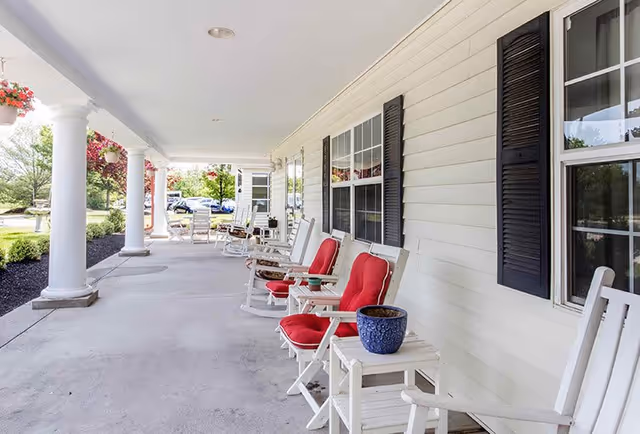 A spacious covered porch at Voorhees Senior Living with white rocking chairs featuring red cushions, small white tables, potted plants, and hanging flower baskets. The porch overlooks a landscaped garden area with trees and shrubs.