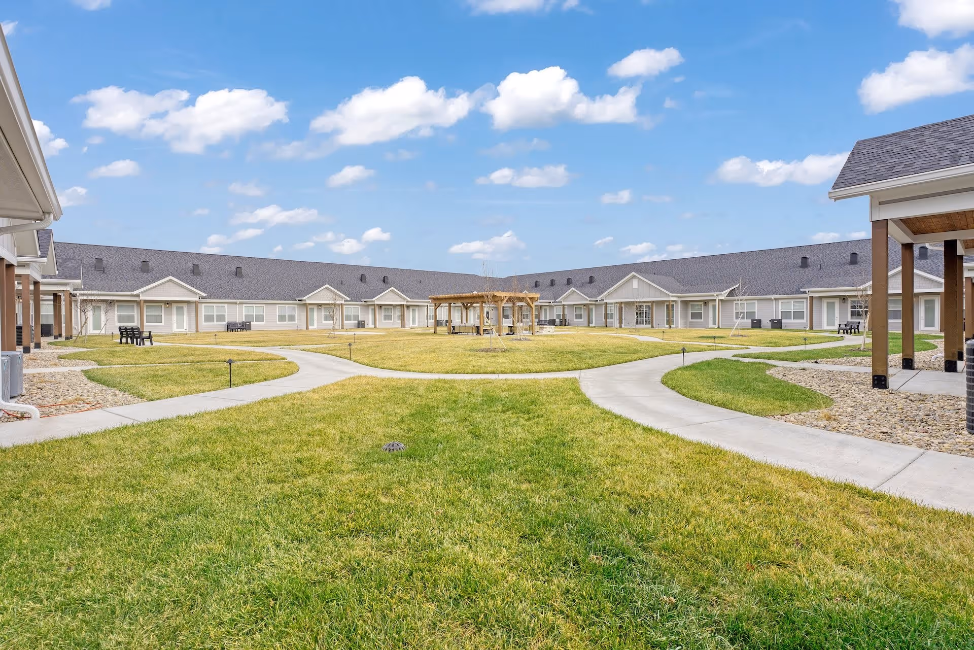 Outdoor courtyard area of Cedarhurst Senior Living of Salina featuring a large grassy lawn with paved walkways, a wooden pergola in the center, and single-story residential buildings surrounding the courtyard under a partly cloudy blue sky.