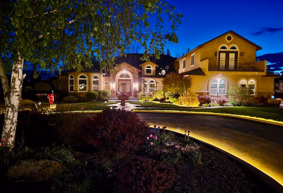 Large illuminated two-story house with a curved lit driveway and landscaped front yard at dusk.