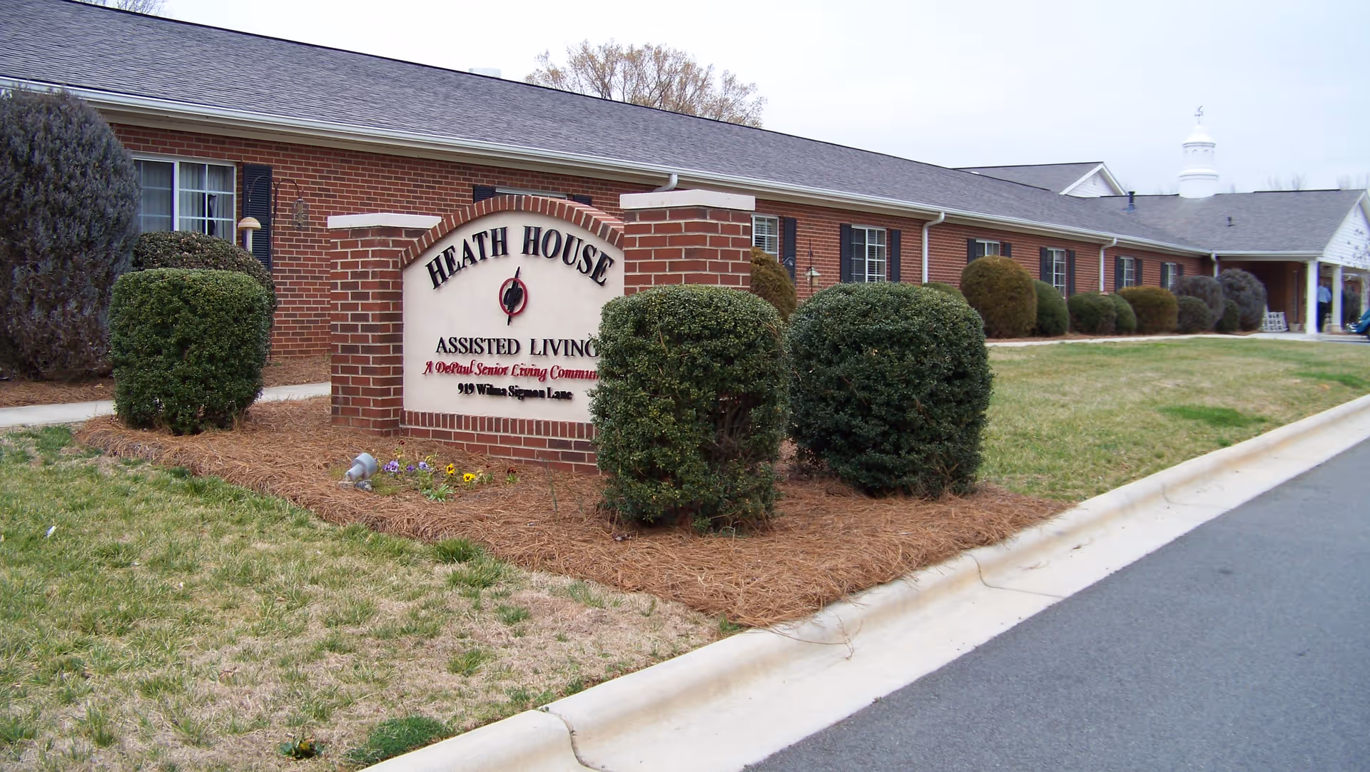 Exterior view of Heath House Assisted Living facility showing a brick building with multiple windows and neatly trimmed bushes in front. A large brick sign with the facility name and address is prominently displayed near the sidewalk.