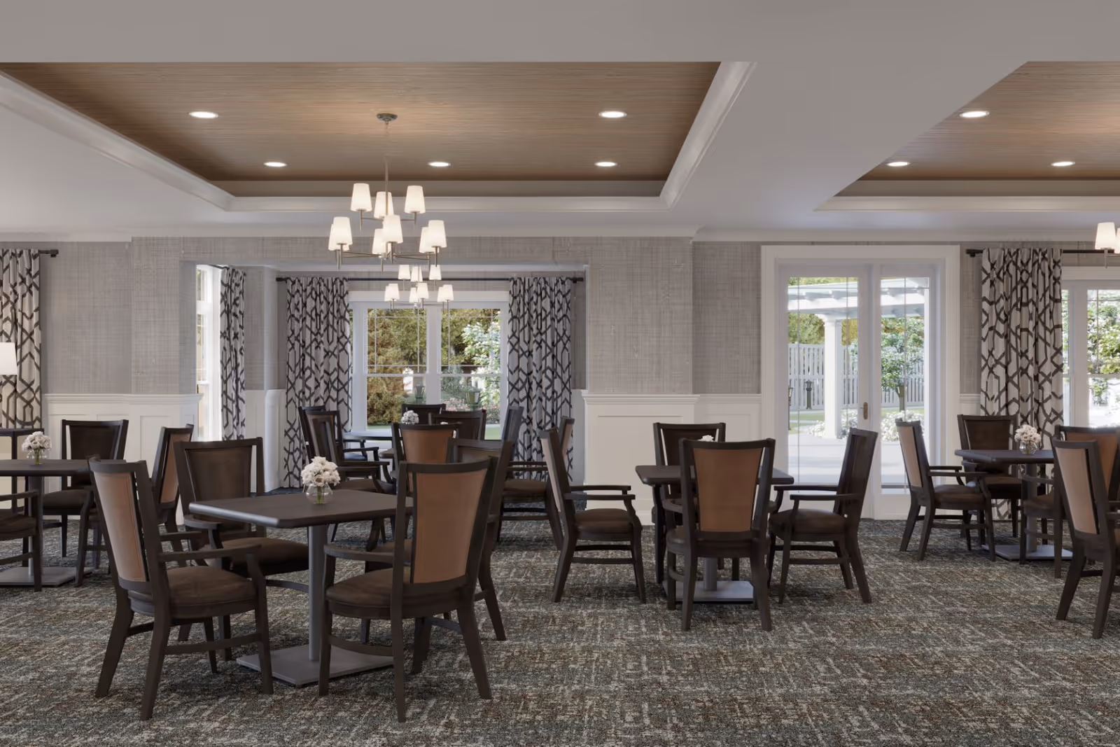 A well-lit dining room with multiple square tables and wooden chairs arranged neatly. Each table has a small vase with white flowers. The room features patterned curtains on large windows and glass doors that open to an outdoor area. The ceiling has recessed lighting and two modern chandeliers.