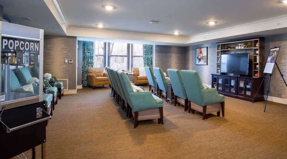 A cozy common area with rows of teal upholstered chairs facing a large flat-screen TV on a dark wooden entertainment center. There are two tan armchairs near a window with blue patterned curtains, a popcorn machine on the left, and a framed colorful abstract painting on the wall.