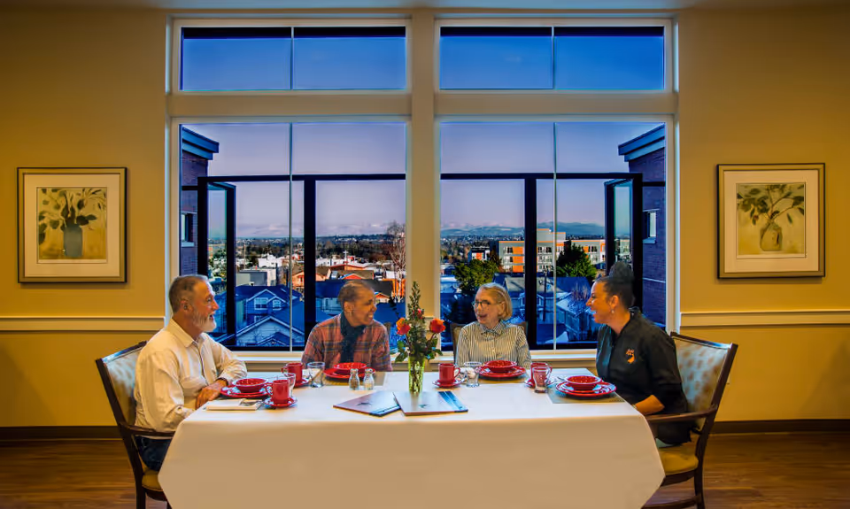 Four people sitting around a dining table set with red plates and cups, engaged in conversation. The table has a white tablecloth and a vase with flowers in the center. Large windows behind them show a cityscape with buildings and mountains in the distance. The room has warm lighting and framed botanical artwork on the walls.