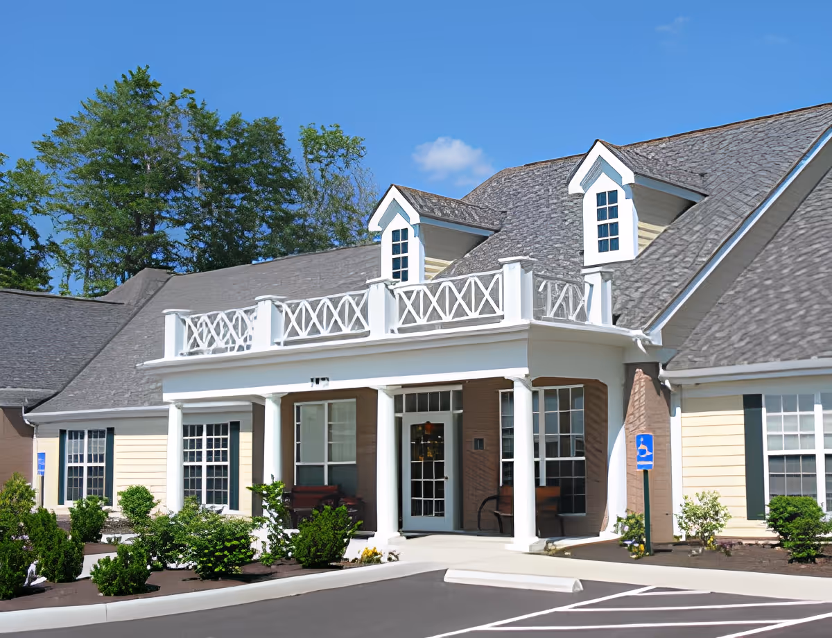 Exterior view of Abbington of Powell Assisted Living facility showing a large building with a gray shingled roof, white columns at the entrance, and beige siding. There are landscaped bushes and plants in front, a clear blue sky, and a handicapped parking sign near the entrance.