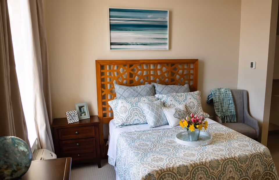A cozy bedroom with a wooden headboard bed adorned with multiple patterned pillows and a matching bedspread. A small wooden nightstand with two framed photos sits to the left of the bed. A gray upholstered chair with a green throw blanket is placed in the corner. Above the bed hangs a framed picture of an ocean scene. Natural light filters in through a window with beige curtains on the left side.