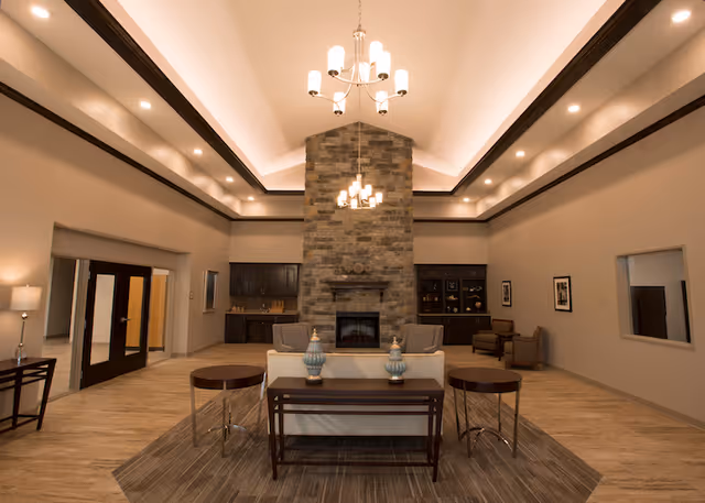 Spacious communal lounge with a central stone fireplace, high tray ceiling with chandeliers, and seating arranged around a rug.