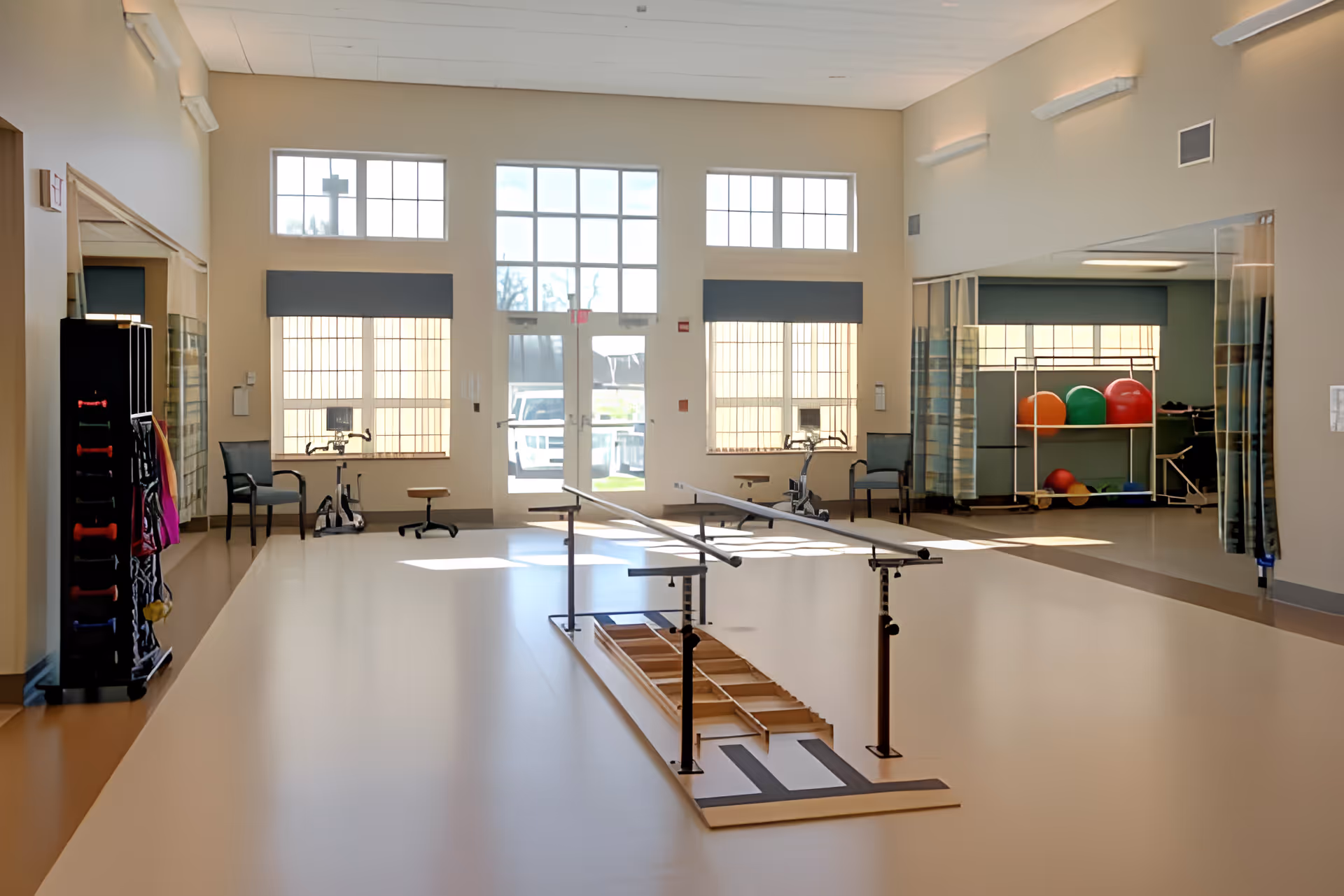 Bright and spacious rehabilitation room with parallel bars for walking exercises in the center, exercise bikes and chairs near large windows, and colorful exercise balls on a rack against the wall.
