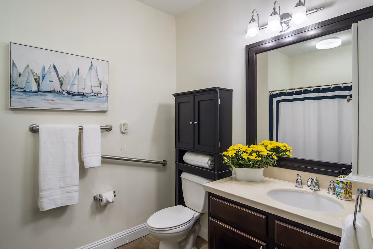 A clean and well-lit bathroom featuring a toilet with a black cabinet above it holding a rolled towel, a sink with a beige countertop and dark wood cabinets below, a large mirror with a dark frame above the sink, a vase of yellow flowers on the countertop, a towel rack with two white towels, a toilet paper holder, a shower curtain with a blue and white border, and a painting of sailboats on the wall.