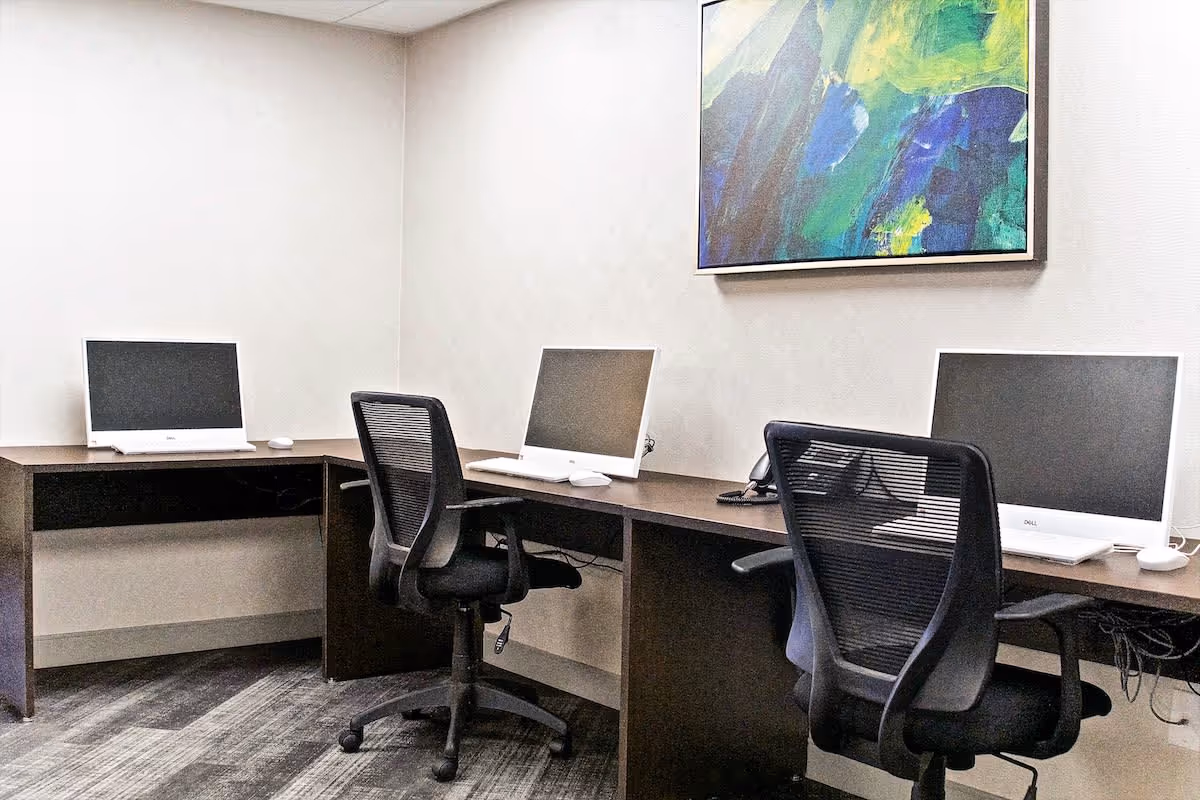 A small office or computer room with three desktop computers on a dark wooden desk arranged in an L-shape. Two black mesh office chairs are positioned in front of the computers. A colorful abstract painting hangs on the white wall above the desk.