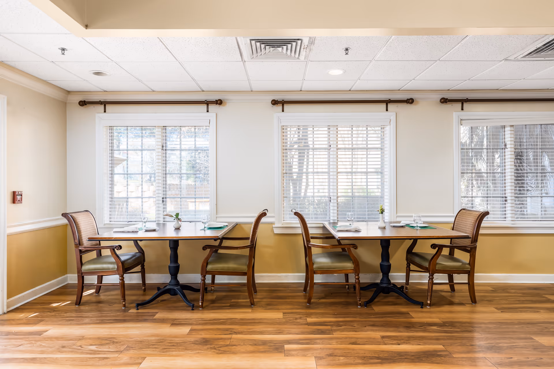 Bright dining area with two wooden tables and four chairs set in front of three large windows.