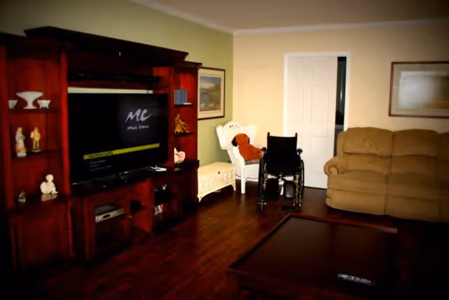 A cozy living room with wooden flooring featuring a large dark wood entertainment center with a flat-screen TV, a beige sofa, a white chair with a teddy bear on it, a wheelchair, and a wooden coffee table with a remote control on it. The walls are painted in light colors with framed artwork hanging.