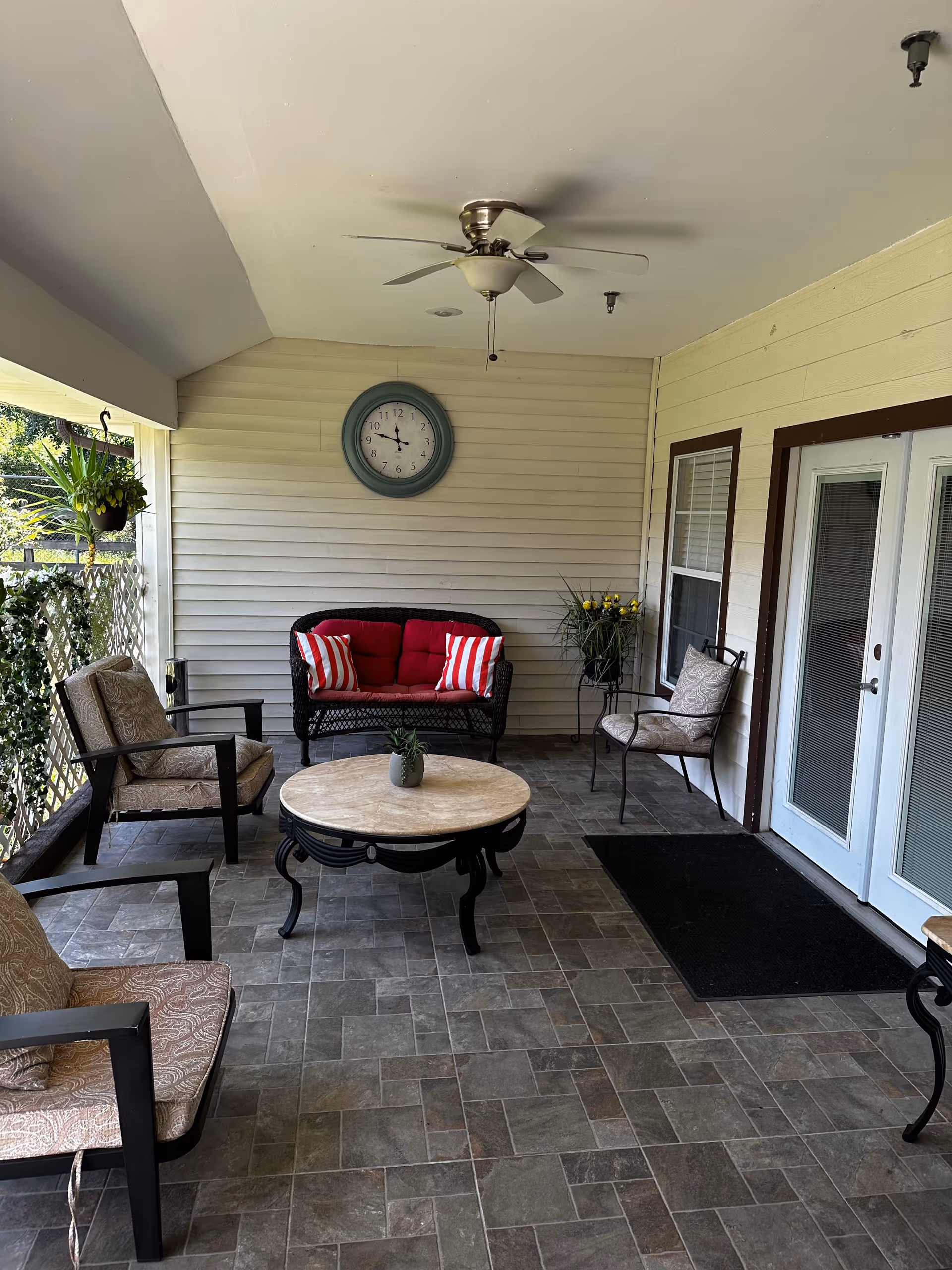 Covered outdoor patio seating area with chairs, a loveseat with red cushions, round coffee table, ceiling fan, and wall clock.