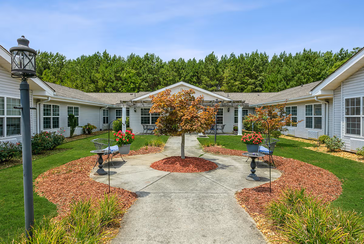Outdoor courtyard area at Seven Hills Square featuring a circular concrete pathway with a small tree in the center, surrounded by mulch and flower pots with red flowers. The courtyard is bordered by a single-story building with white siding and multiple windows, and there is a lamppost on the left side. In the background, there is a dense line of tall green trees under a clear blue sky.