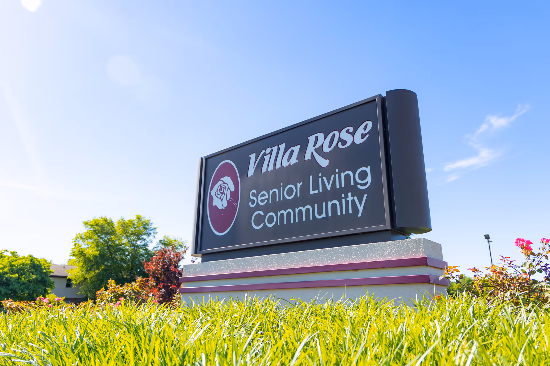 Outdoor sign reading "Villa Rose Senior Living Community" on a landscaped lawn under a blue sky.