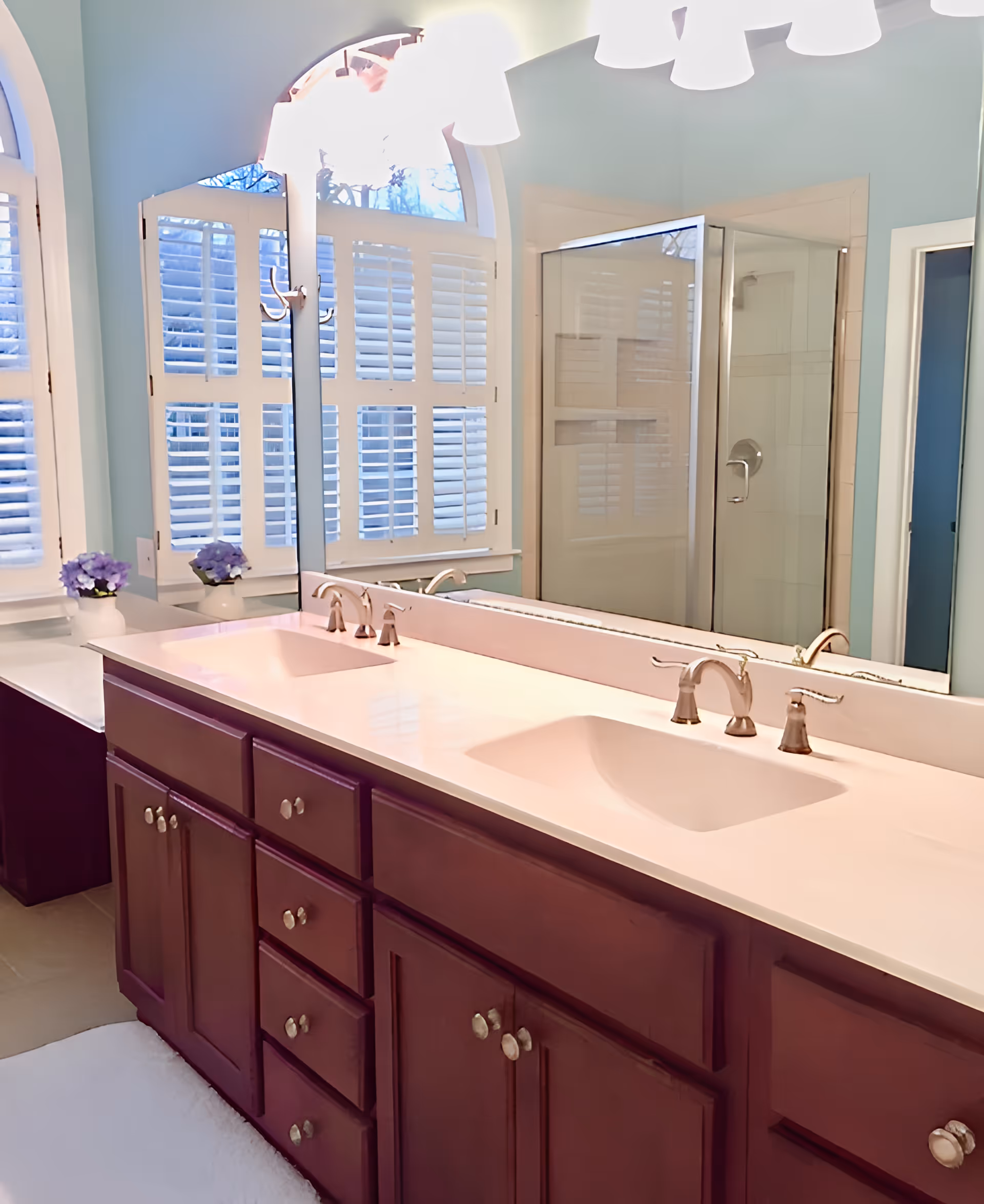 A bathroom with a double sink vanity featuring dark wood cabinets and white countertops. Above the sinks is a large mirror with multiple light fixtures. In the background, there is a glass-enclosed shower and windows with white plantation shutters. A small vase with purple flowers is placed on the countertop near the window.