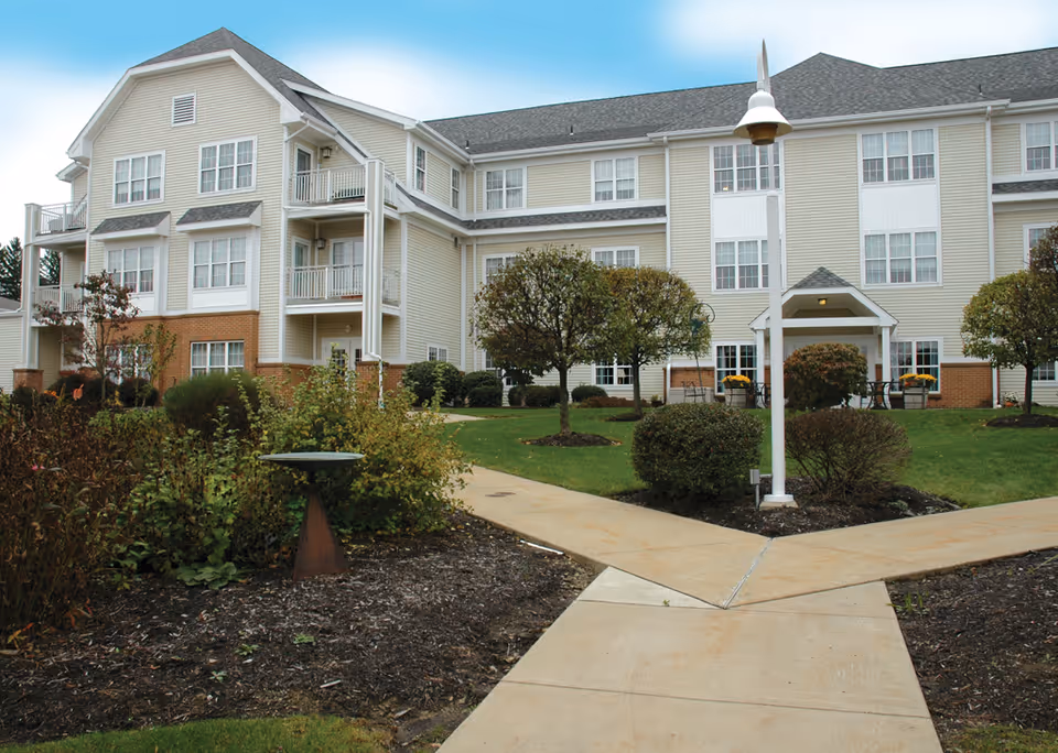 Exterior view of a multi-story senior living facility with beige siding and brick accents. The building is surrounded by a well-maintained garden with bushes, small trees, and a paved walkway leading to the entrance. A white lamp post is visible near the walkway.