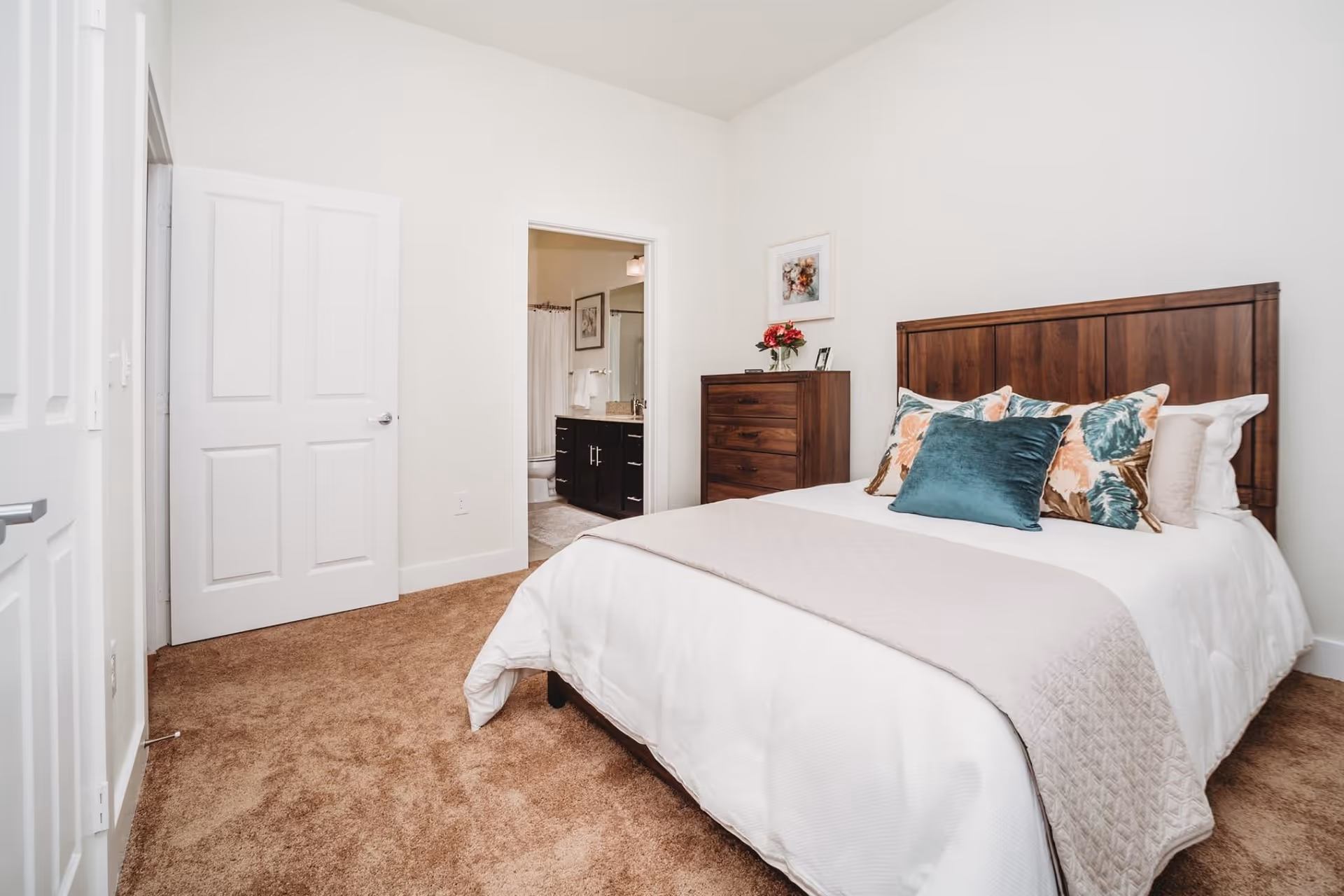 A bedroom with a wooden headboard bed dressed in white bedding and decorative pillows in teal and floral patterns. There is a wooden dresser with a vase of flowers and a framed picture above it. The room has beige carpet and white walls. An open door reveals a bathroom with a vanity and mirror.