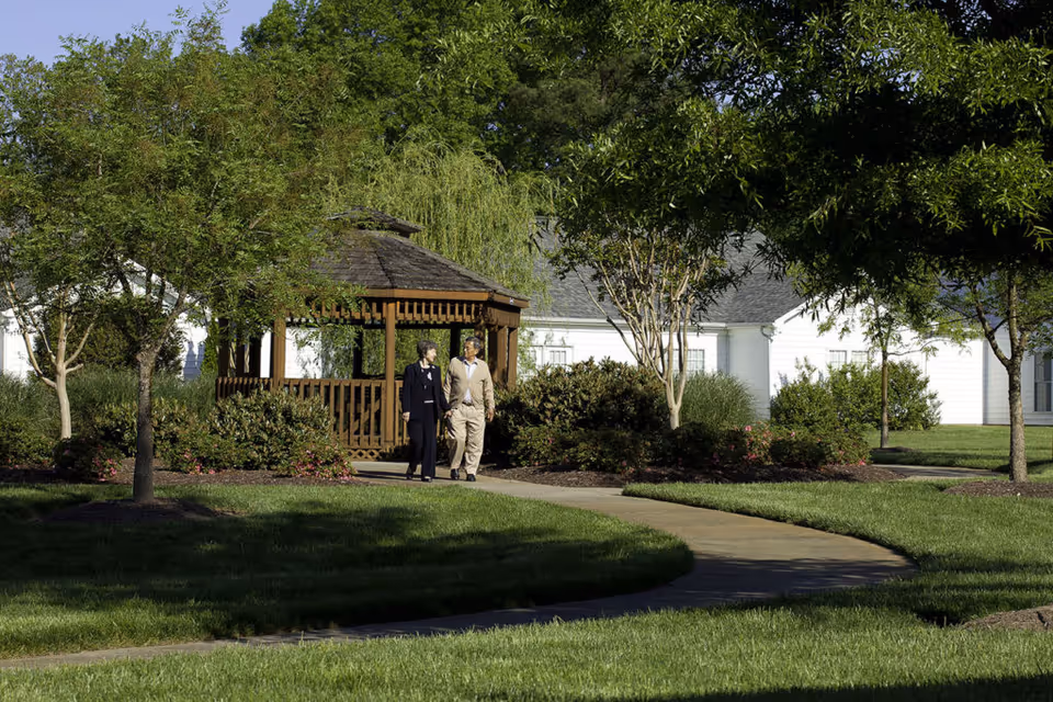 Two elderly people walking on a curved paved path in a well-maintained garden area with green grass, trees, and bushes. A wooden gazebo is visible in the background along with white buildings under a clear sky.