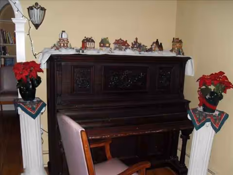 Dark wooden piano topped with miniature holiday village figures, poinsettias, and a pink chair