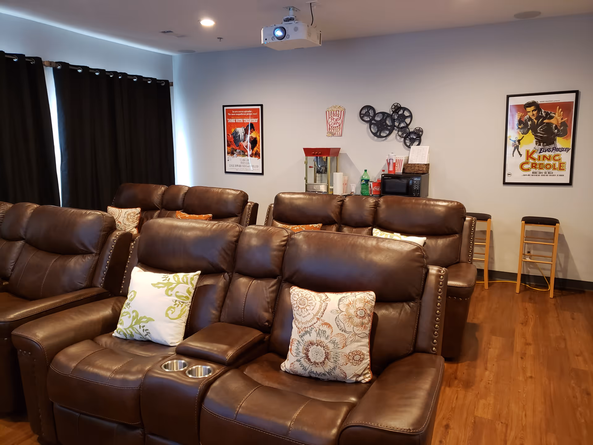 A cozy movie room with several brown leather recliner chairs arranged in rows, each with decorative pillows. On the back wall, there is a popcorn machine, a microwave, soda bottles, and two movie posters. The room has wooden flooring and dark curtains covering the windows.