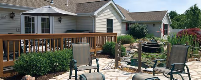 Outdoor patio area at The Parkvue Community On Catawba Island featuring a stone-paved seating area with several chairs, a wooden railing, a small pond with rocks and plants, and a light-colored building with windows and a door in the background.