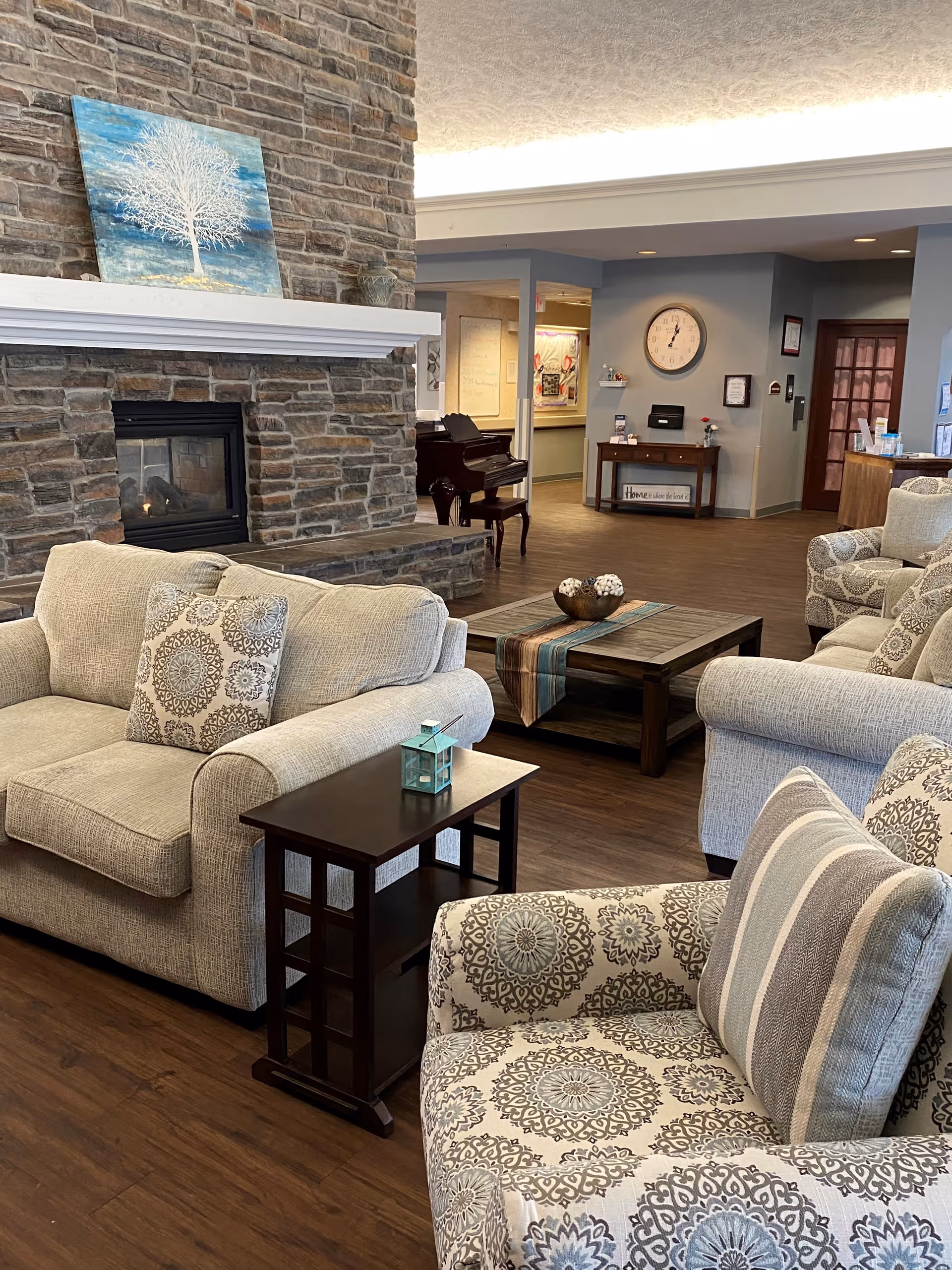 A cozy living room area with beige and patterned armchairs and sofas arranged around a wooden coffee table with a decorative runner and bowl. There is a stone fireplace with a painting of a white tree on a blue background above it. In the background, a piano is visible along with a wall clock and a small table with framed items and a sign that reads 'Home is where the heart is.' The room has wooden flooring and soft lighting.