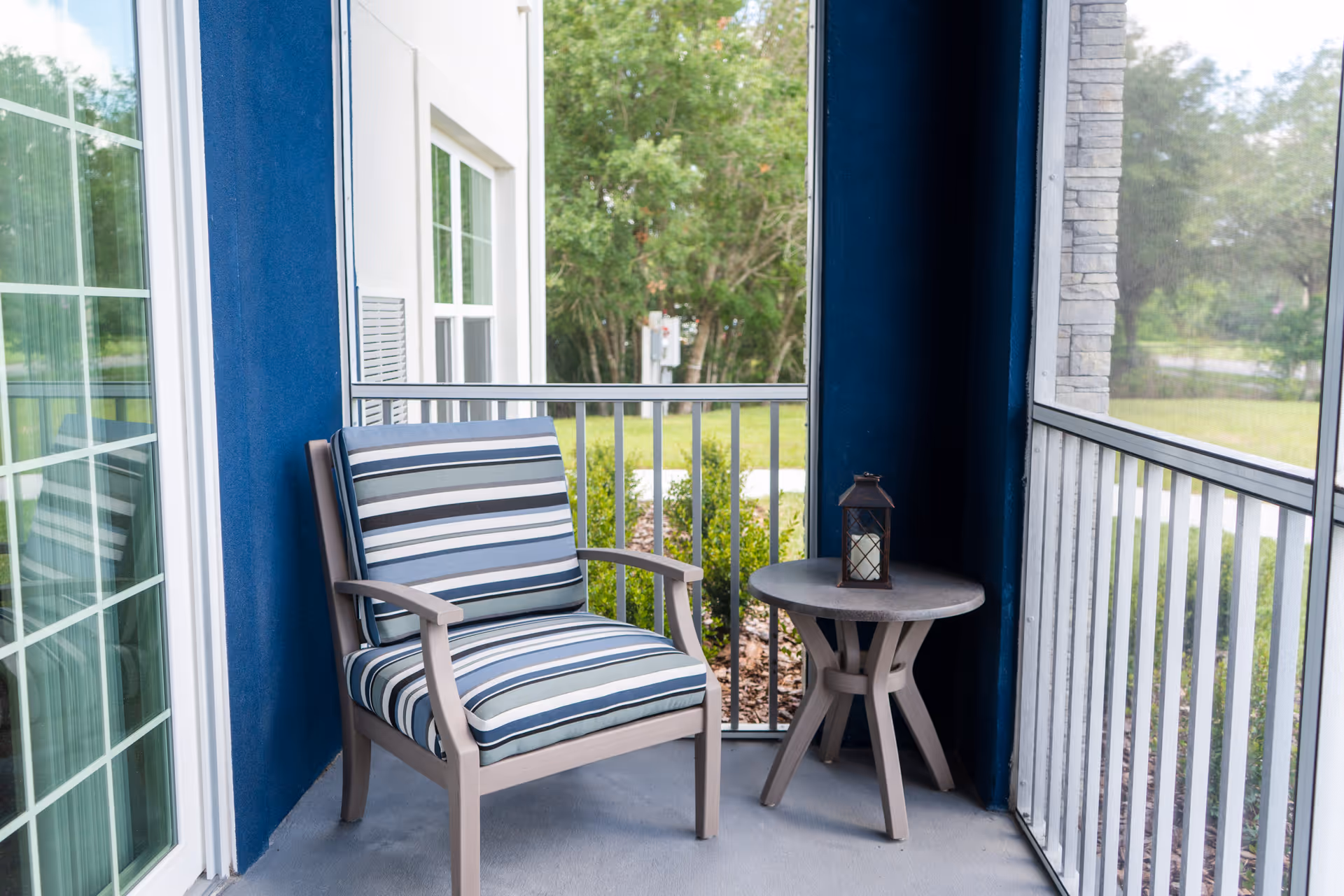 A small screened-in porch with a cushioned armchair featuring blue, white, and gray stripes, and a round side table with a decorative lantern on top. The porch overlooks a green outdoor area with trees and bushes.