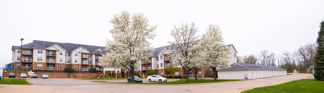 Exterior view of Pennsylvania Place AL, a multi-story senior living facility with balconies, surrounded by blooming trees and a parking area with several cars.