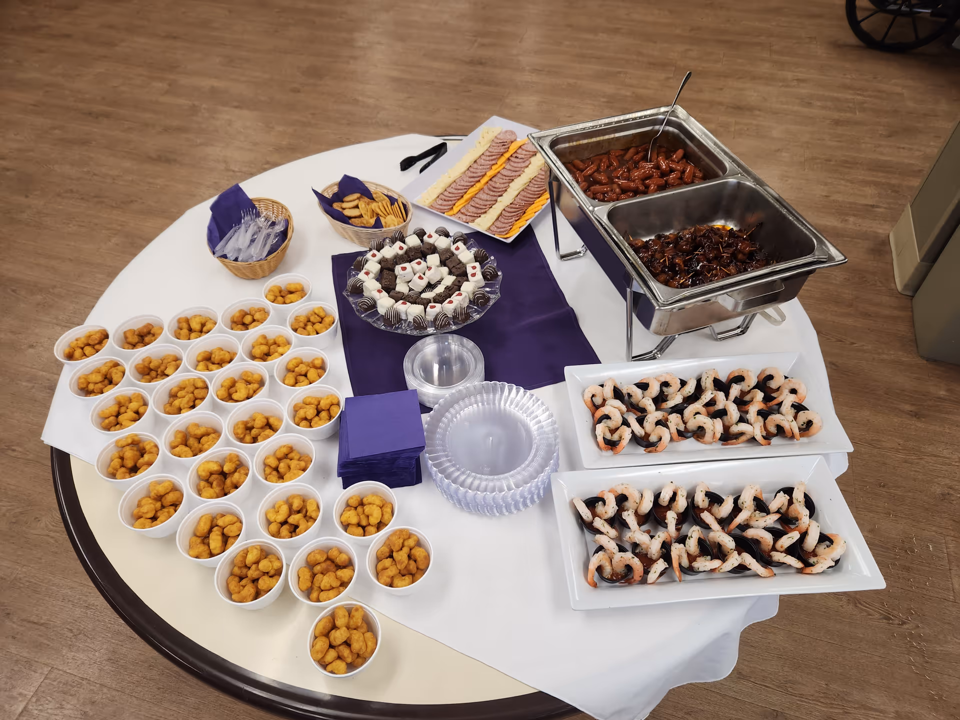 A round table covered with a white tablecloth and a purple runner, displaying a variety of appetizers including cups filled with small fried snacks, trays of shrimp on seaweed, a platter of sliced meats and cheeses, a plate of small desserts, and two metal chafing dishes containing cooked sausages and another meat dish. There are also plastic utensils, napkins, and clear plastic plates on the table.