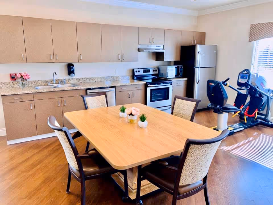 A bright kitchen and dining area with wooden cabinets, granite countertops, a stainless steel sink, stove, microwave, and refrigerator. A wooden dining table with four cushioned chairs is centered in the room, decorated with small potted plants and flowers. In the background, there is exercise equipment near a window with blinds letting in natural light.