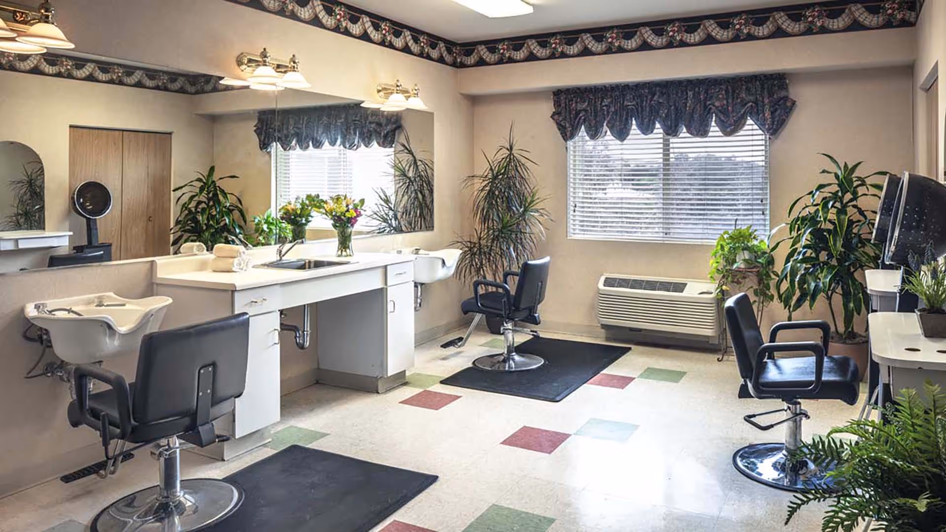 Interior of a hair salon area with three black salon chairs, two white hair washing sinks, a large mirror on the wall, plants, and a window with blinds and a decorative valance.