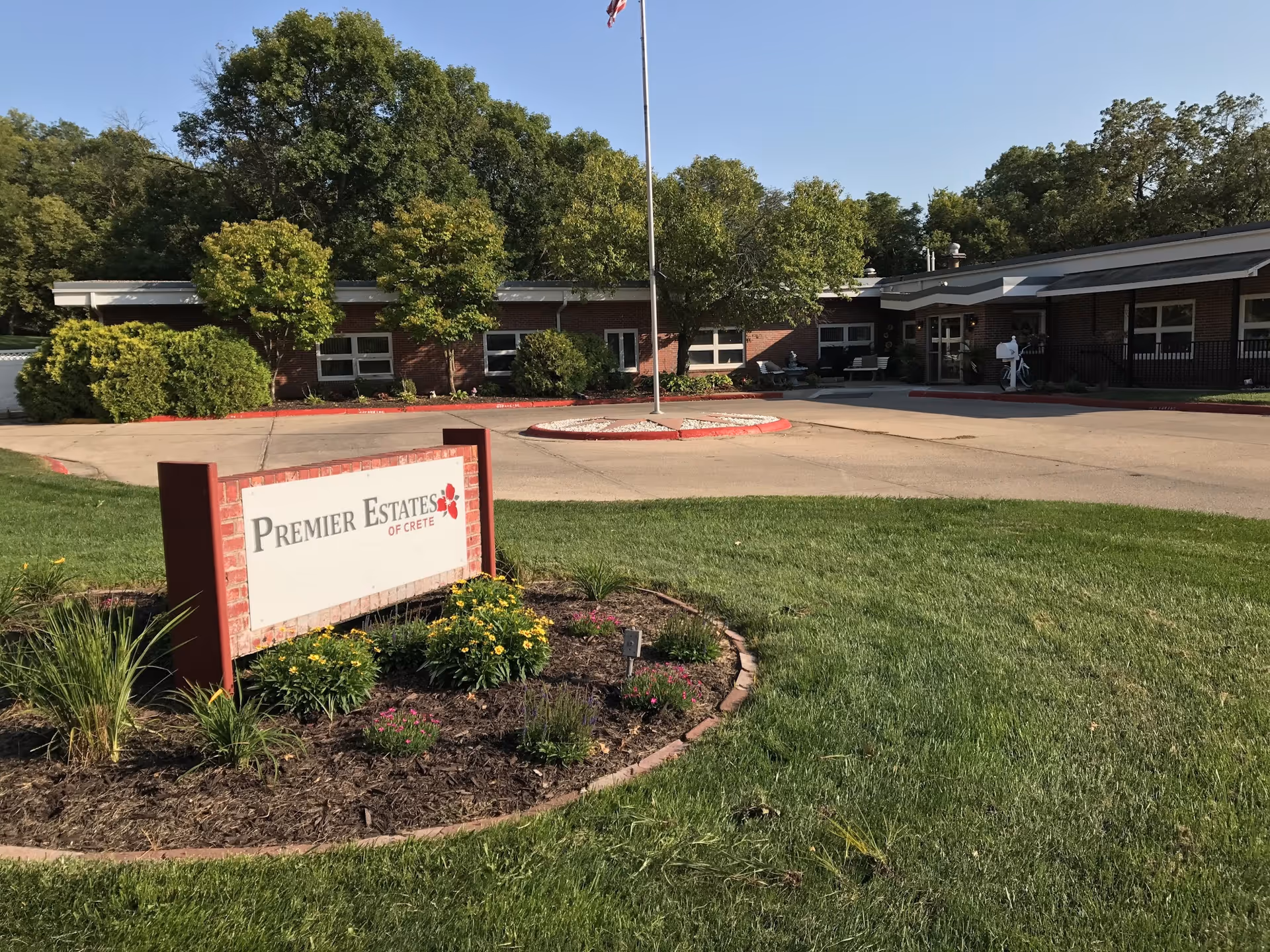 Exterior view of Premier Estates Of Crete, showing a single-story brick building with several windows, a driveway with a circular island featuring a flagpole, and a landscaped area with green grass, bushes, and flowers in front of a sign displaying the facility's name.